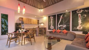 Una sala de estar moderna con una mesa de centro de vidrio, sofá gris con cojines naranjas, sillas de madera y una zona de comedor con mesa y sillas. Hay pinturas abstractas en la pared y una cocina al fondo.