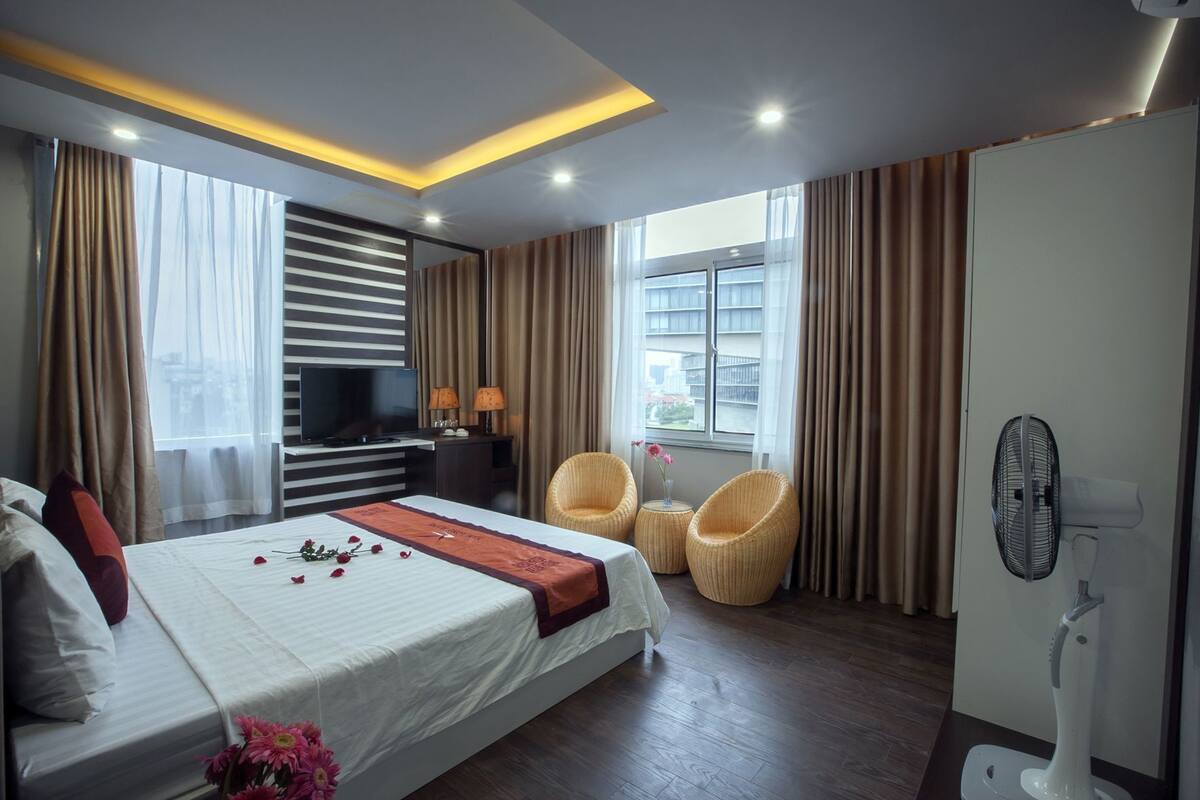 A hotel room with a bed, a television, a desk with lamps, two wicker chairs, a fan, and a window with curtains.