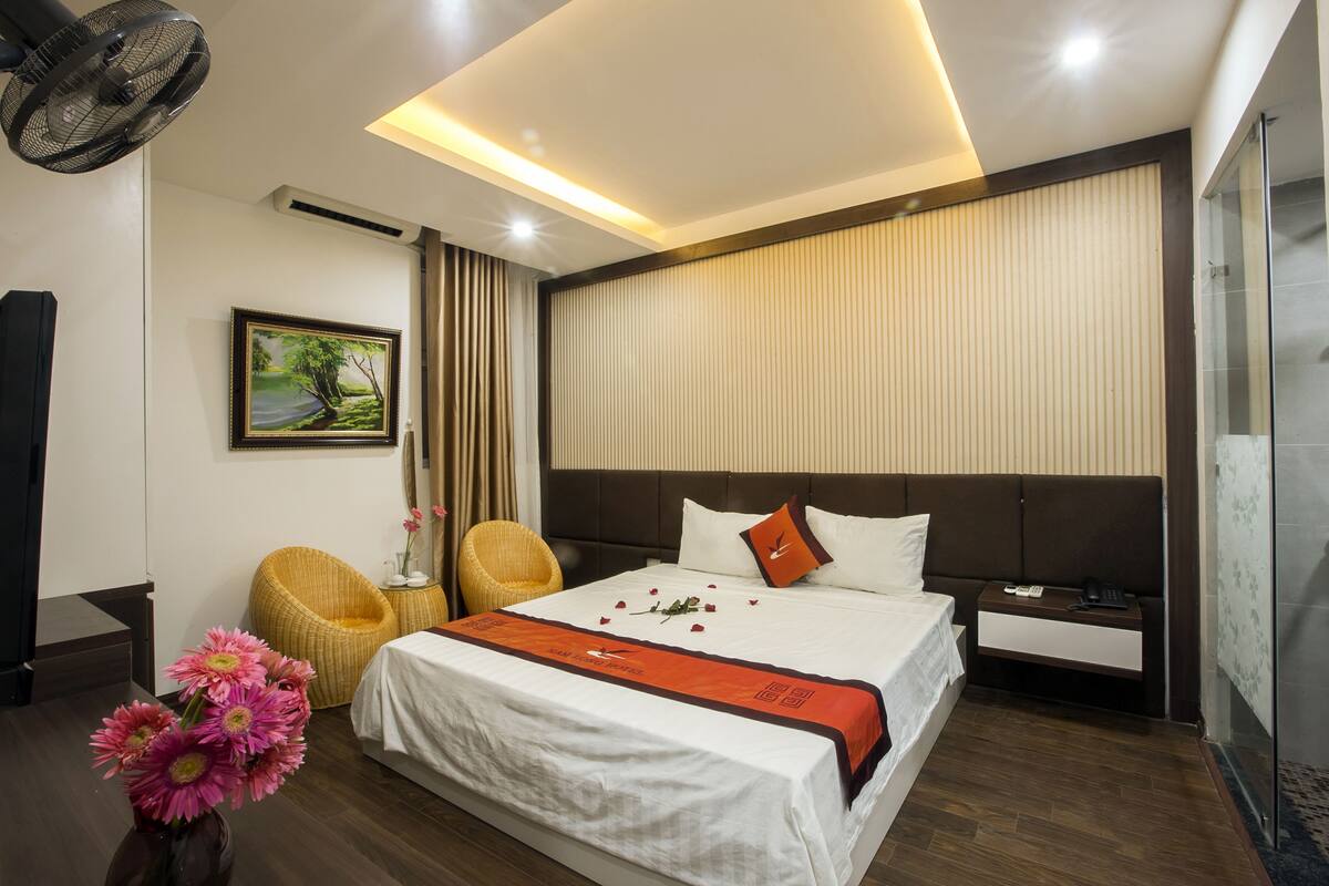 A modern hotel room with a large bed, a bedside table with a phone, a chair, a painting on the wall, and a fan.
