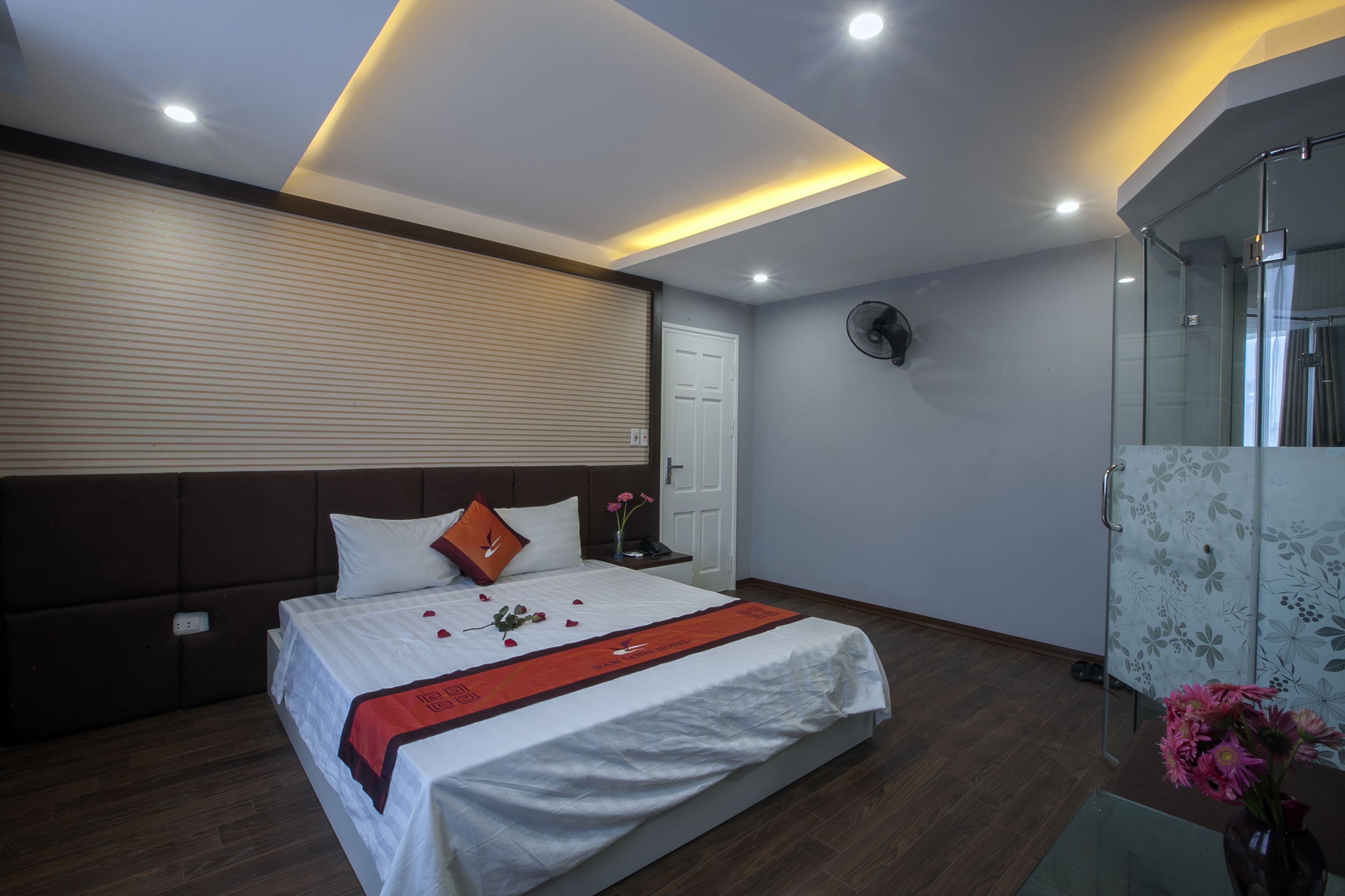 A modern hotel room with a large bed, a bedside table, a wall-mounted fan, and a glass shower enclosure.