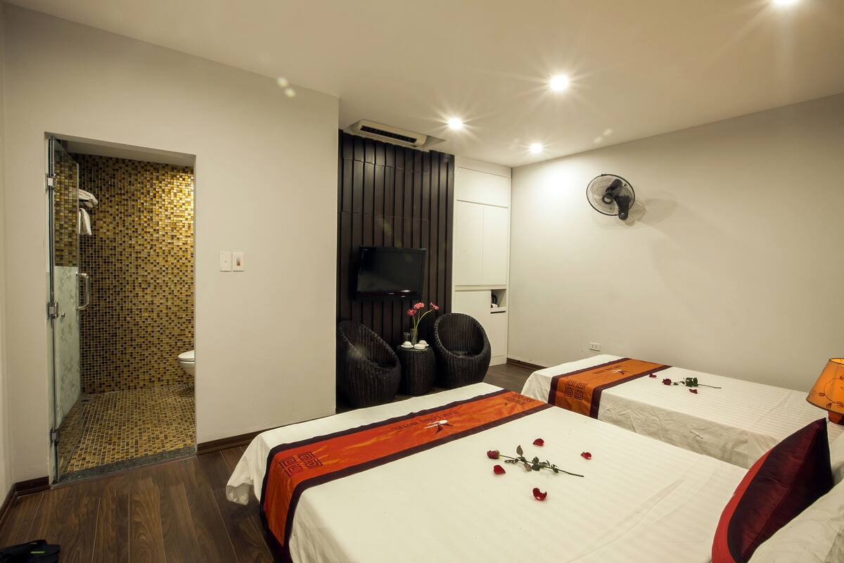 A hotel room with two beds, a shower area, a TV, and a fan.