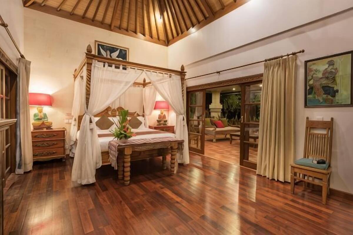 Uma cama com dossel e cortina, piso de madeira, mesa de cabeceira com abajur e uma cadeira.
