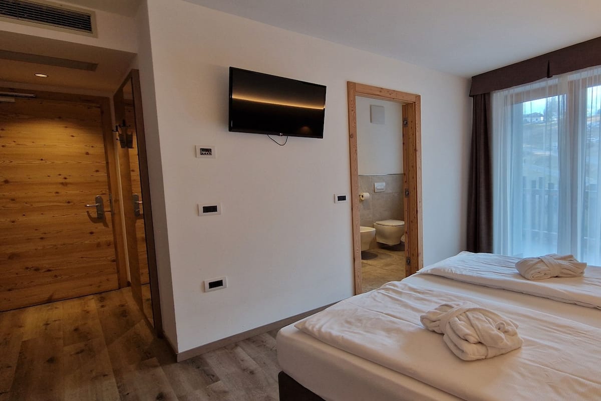 A hotel room with a wooden wardrobe, a television mounted on the wall, a large window with curtains, and two beds with towels.