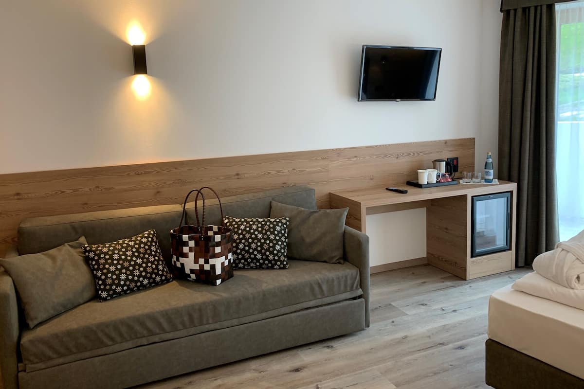 A modern hotel room with a grey sofa, a wooden headboard, a flat-screen TV mounted on the wall, a small wooden desk with a coffee maker, and a mini fridge.