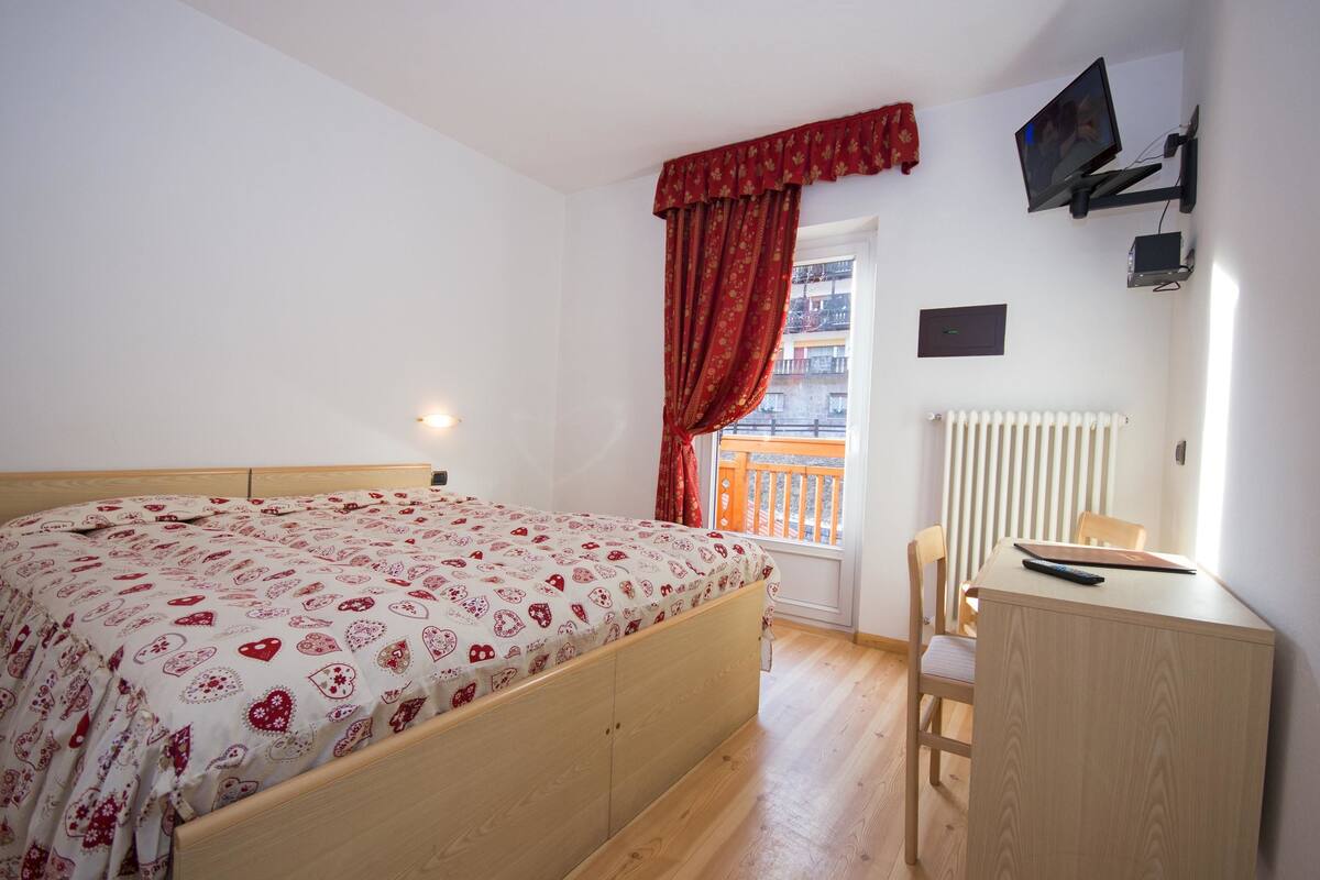 A bedroom with a bed, a desk, a chair, a window with red curtains, and a radiator.