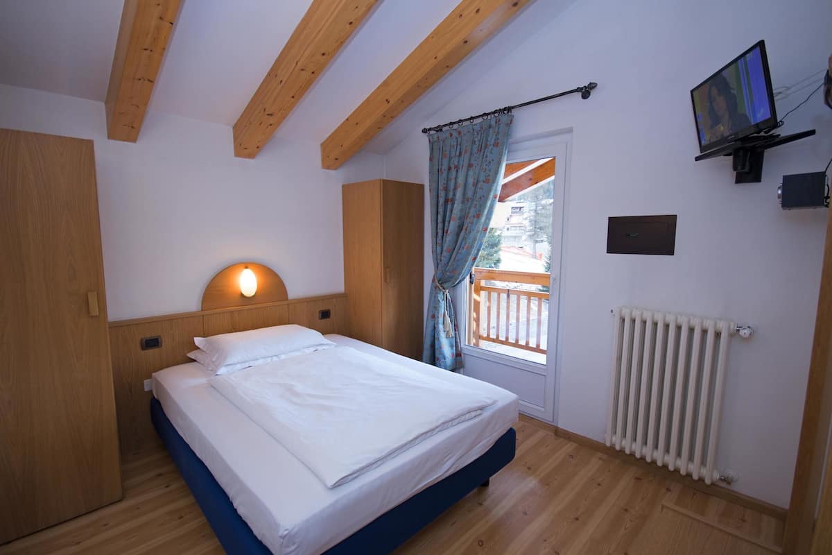 A bedroom with a bed, wooden ceiling beams, a window with curtains, a radiator, and a television on the wall.
