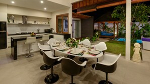 A modern kitchen with a dining area, featuring a round table with white chairs, a central island with bar stools, and a view of a lush garden and pool. - Villa Pacha seminyak by Best deals Asia Hospitality (Seminyak)