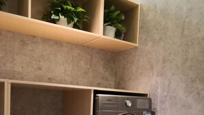 A compact laundry area with a front-loading washer and dryer, wooden shelving with potted plants, and a framed picture on the lower shelf. - ibis Styles Wuhan Optics Valley Square Hotel (Wuhan)