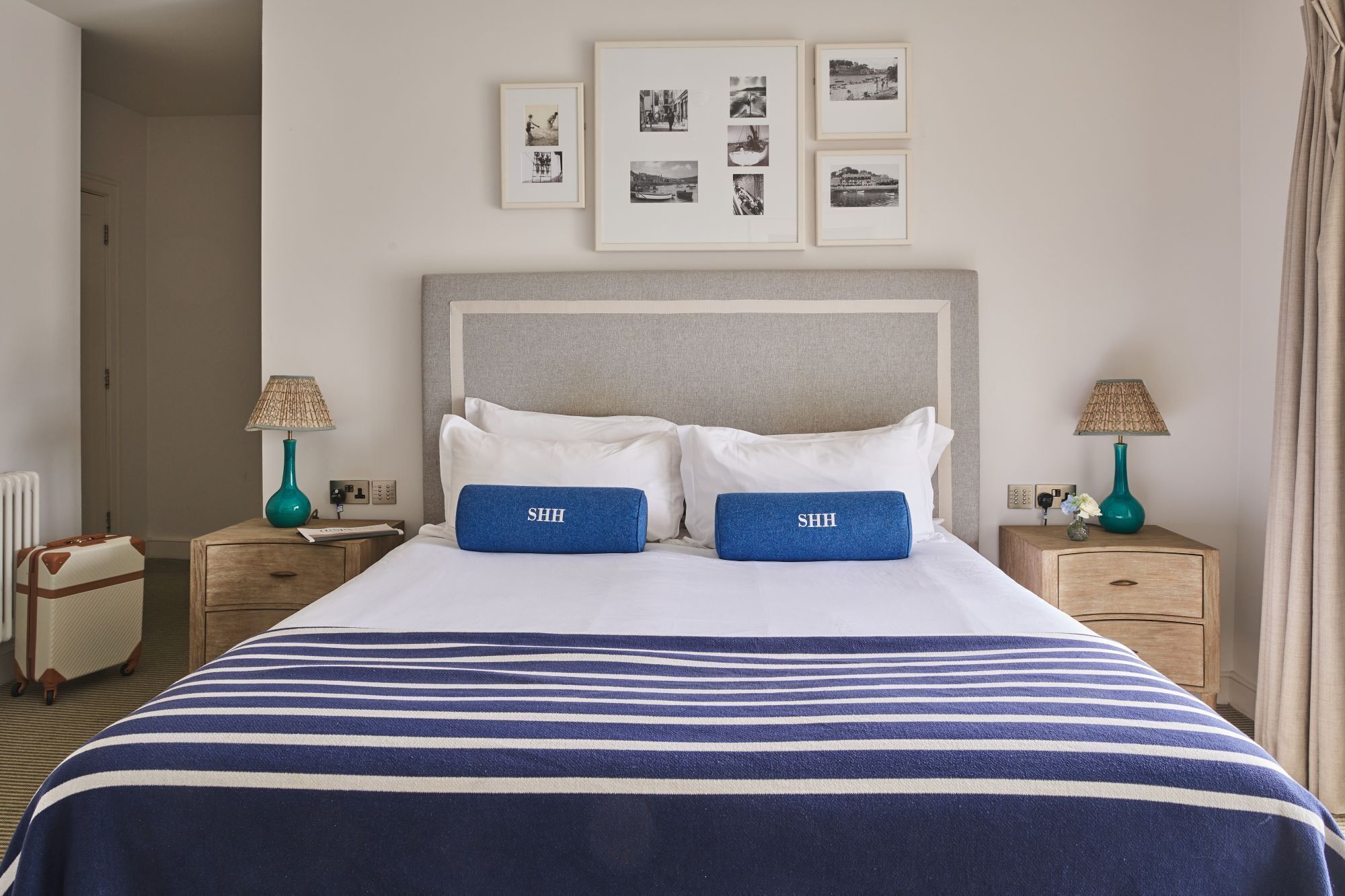 Una cama bien hecha con una colcha a rayas, dos almohadas con la palabra 'SHH' y un cabecero. Encima de la cama hay cuadros enmarcados en la pared.