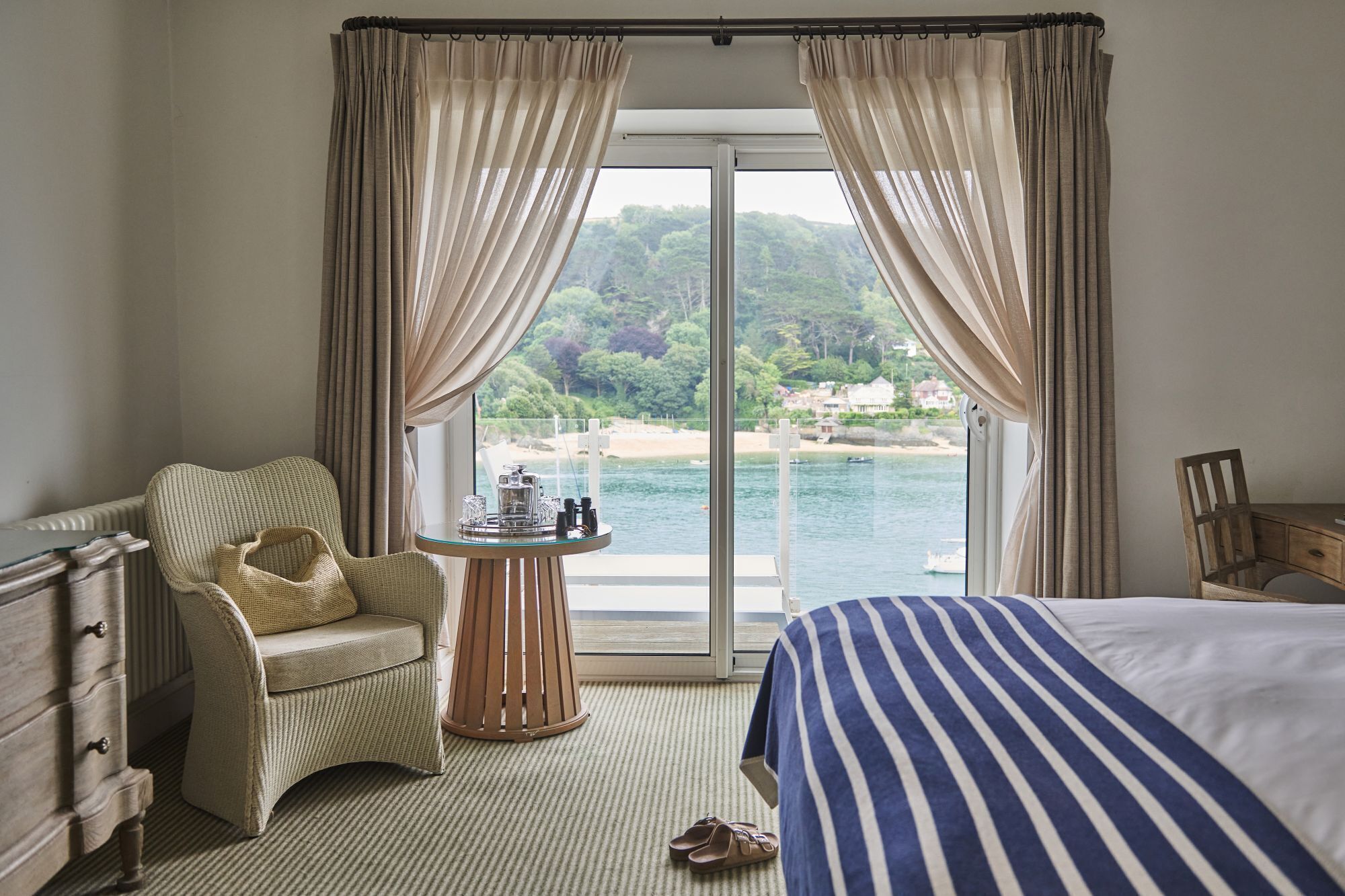 A hotel room with a bed, a chair, a small table with toiletries, and a view of a beach and water.