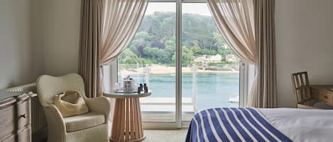 Habitación de hotel con cama, una silla, una mesita con artículos de tocador y vista a una playa y el mar.