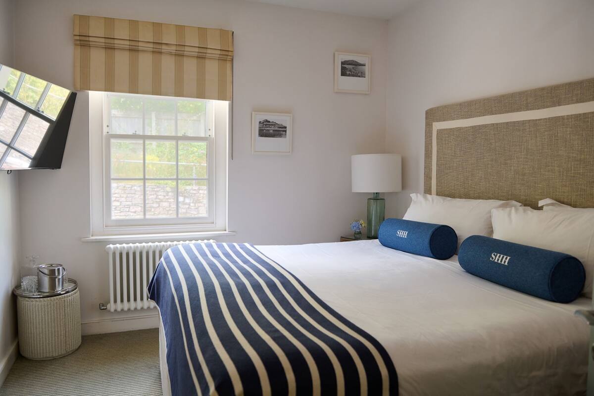 A hotel room with a large bed, a window with blinds, a radiator, a wall-mounted TV, and a small table with a lamp.