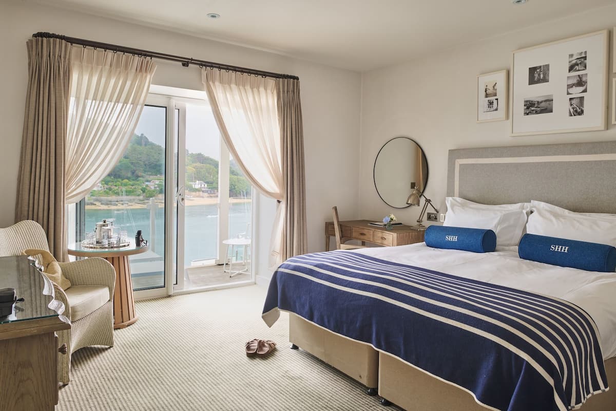 A hotel room with a large bed, a desk with a chair, a round table with a chair, and a view of the sea through the window.