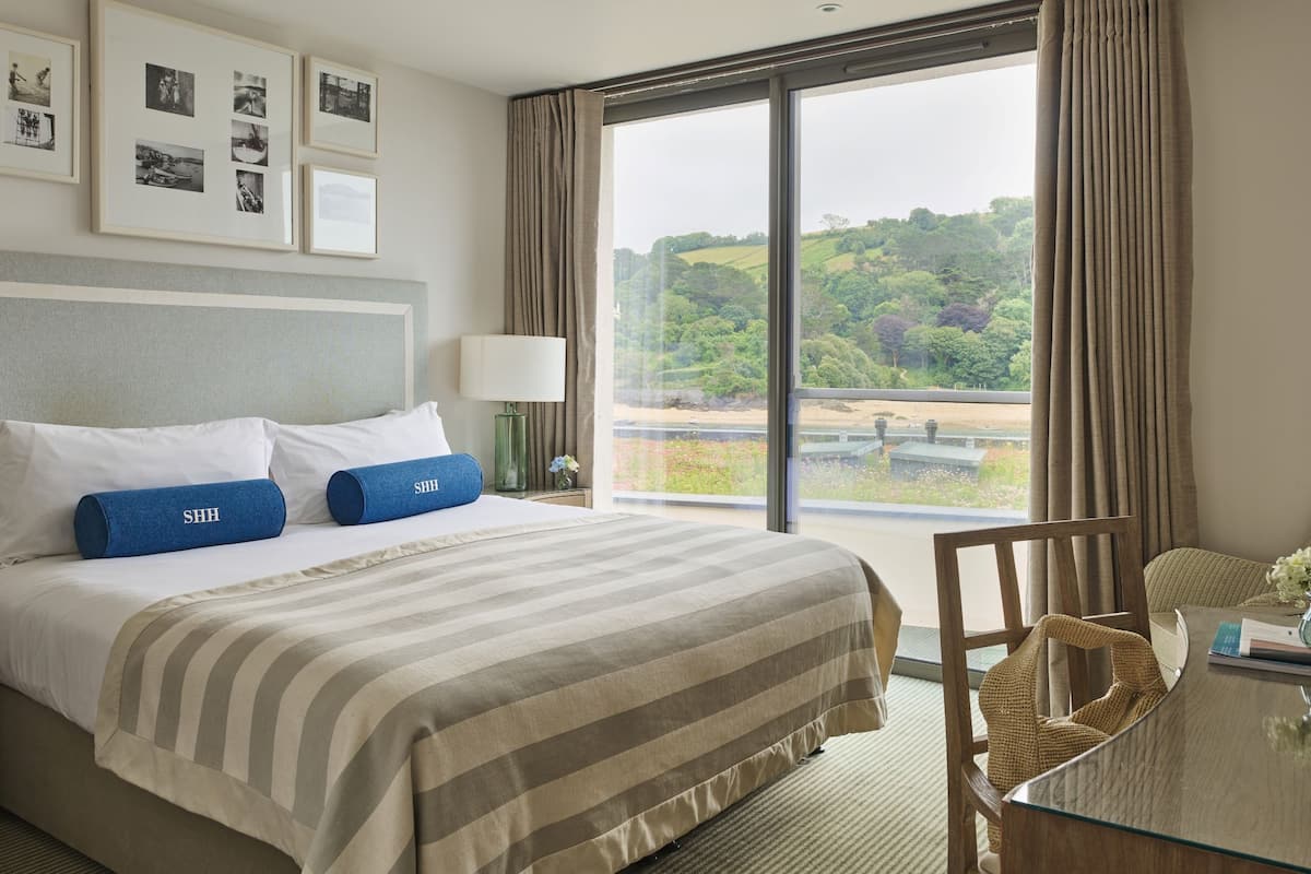 A hotel room with a large bed, a view of greenery, a chair, and a table with a vase.