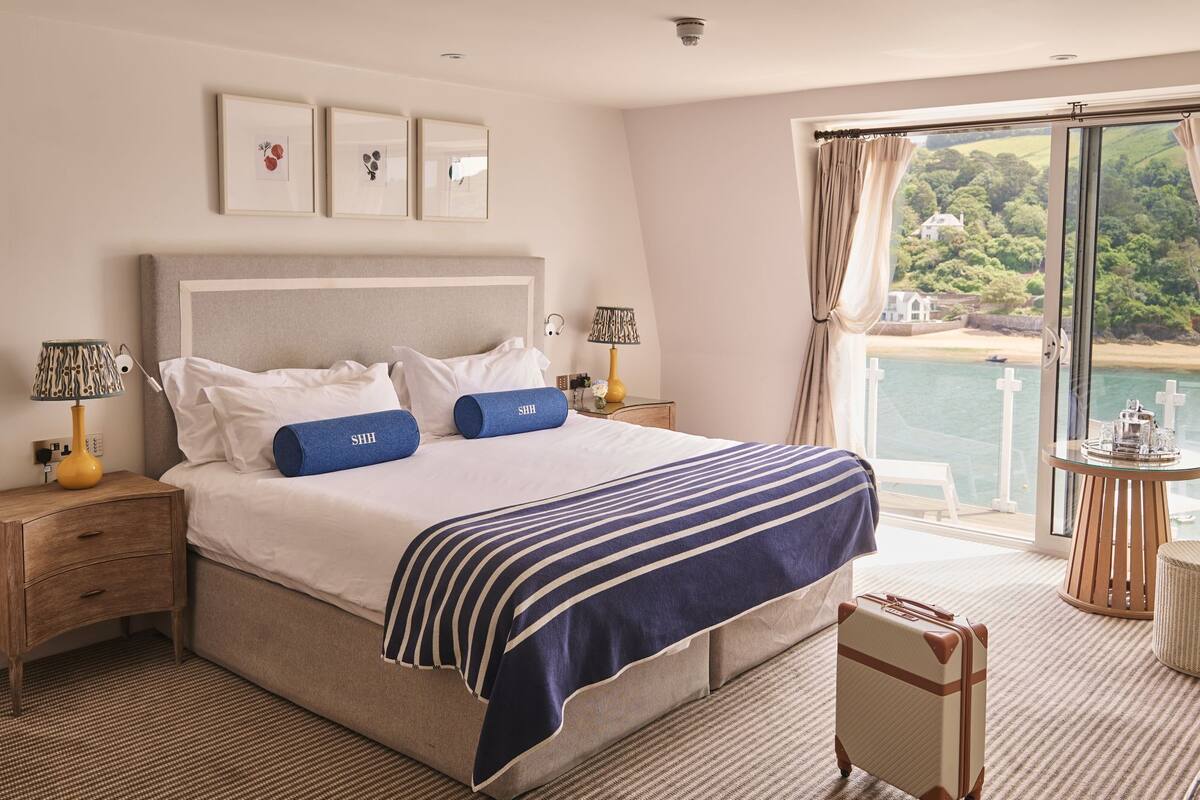 A hotel room with a large bed, two bedside tables with lamps, a view of the sea, and a balcony with a table and chairs.