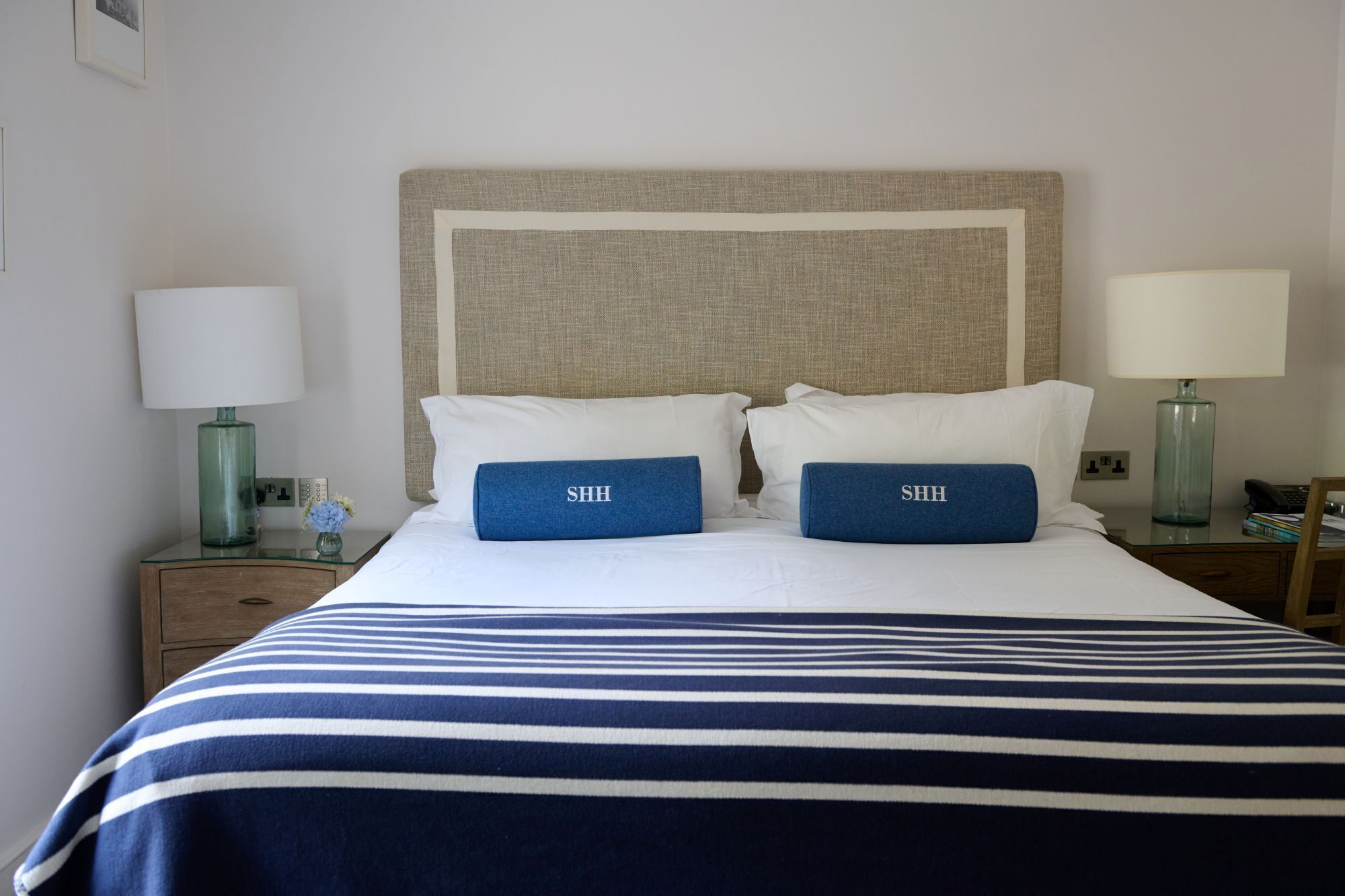 A neatly made bed with a striped bedspread, two pillows with 'SHH' on them, and a headboard with a textured fabric.