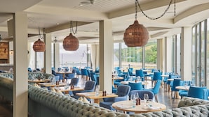 A spacious restaurant with high ceilings, large windows, and a mix of seating including round tables and a long cushioned bench. - Harbour Hotel Salcombe (Salcombe)