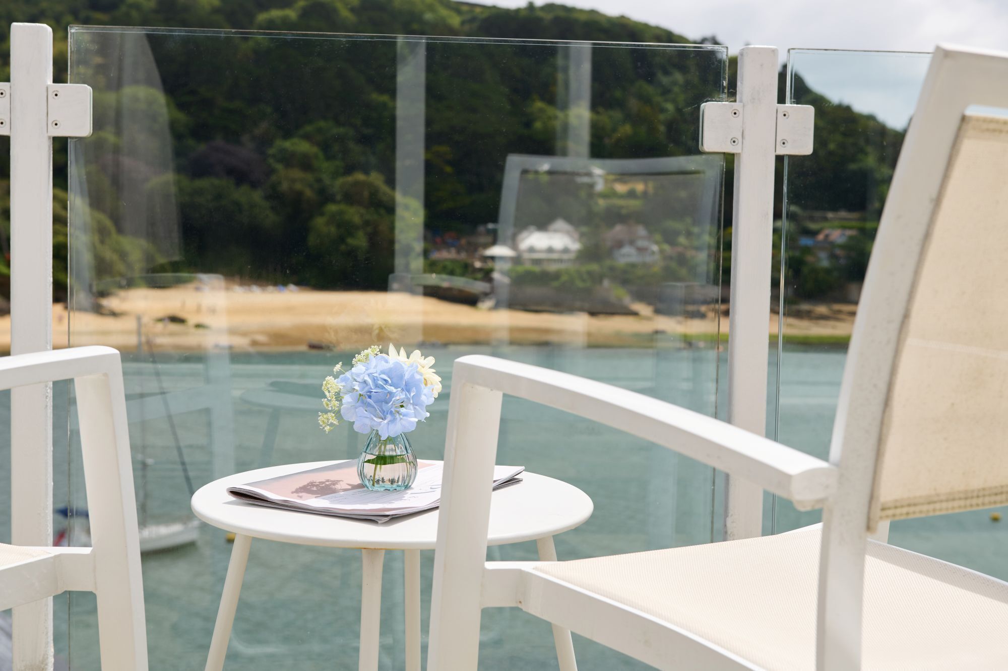 A glass-enclosed balcony with a white chair, a small table with a vase of flowers, and a view of a beach and forested area.