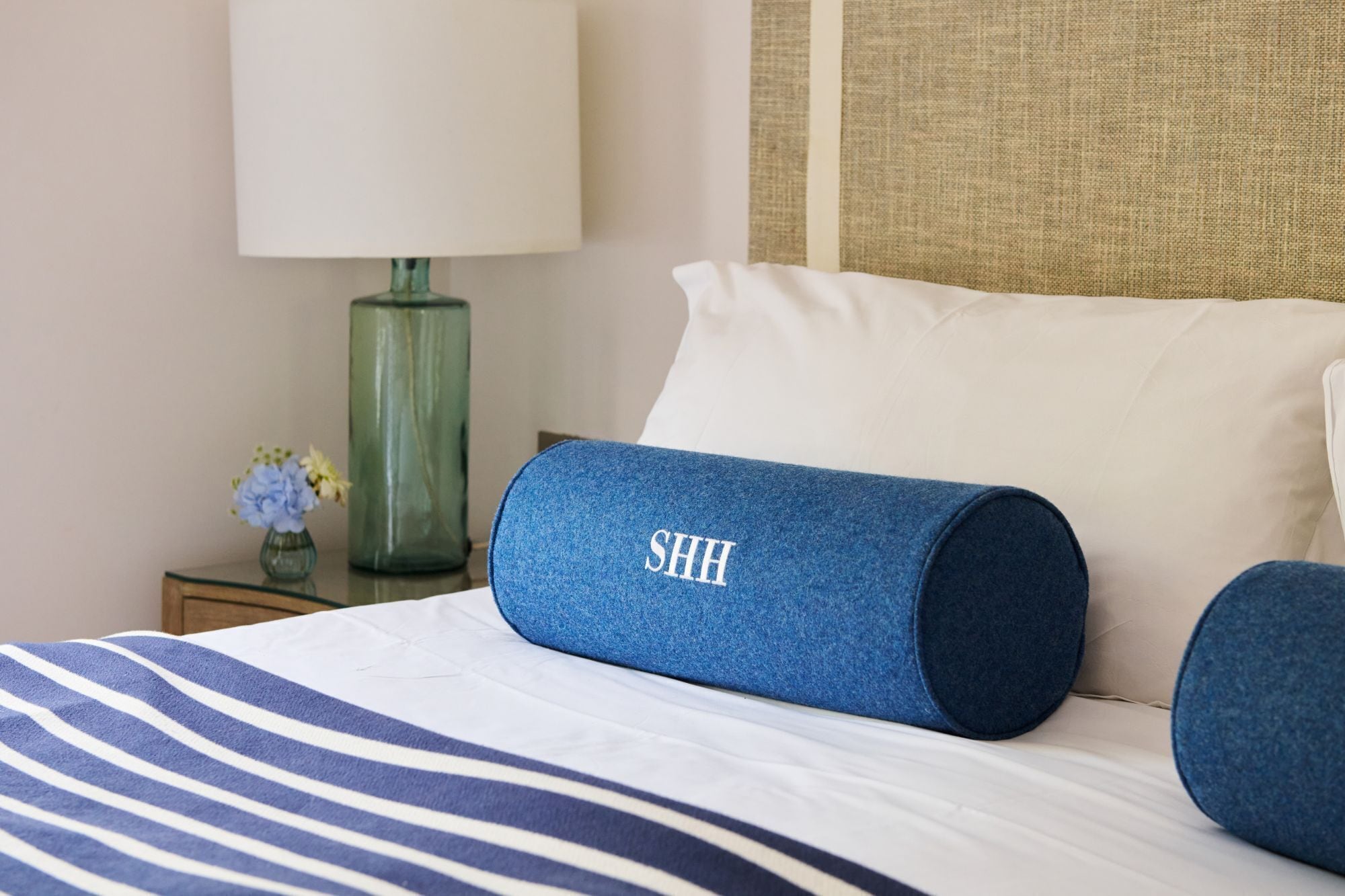 A hotel room with a bed featuring striped bedding, a blue pillow with 'SHH' embroidered, a white pillow, a green vase with flowers, and a lamp on a bedside table.