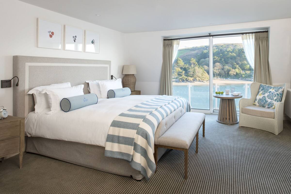 A hotel room with a large bed, a chair, a table with a lamp, and a view of the sea.