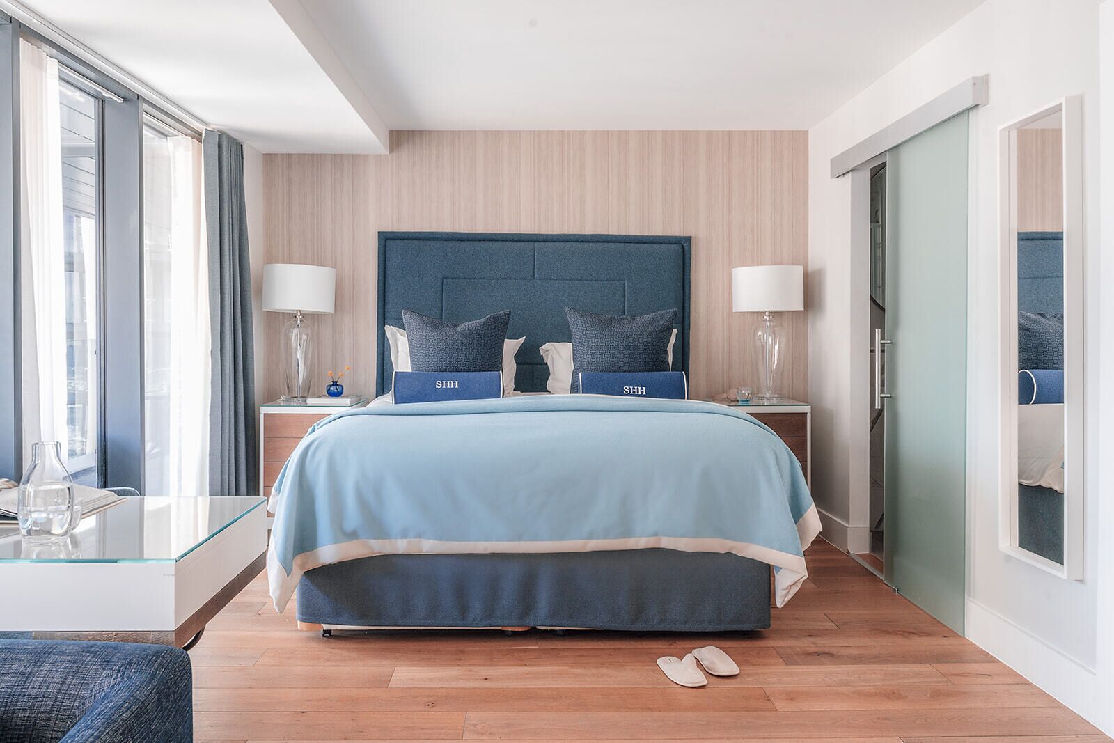 A modern hotel room with a large bed, two bedside tables, a glass coffee table, and a view through a door.