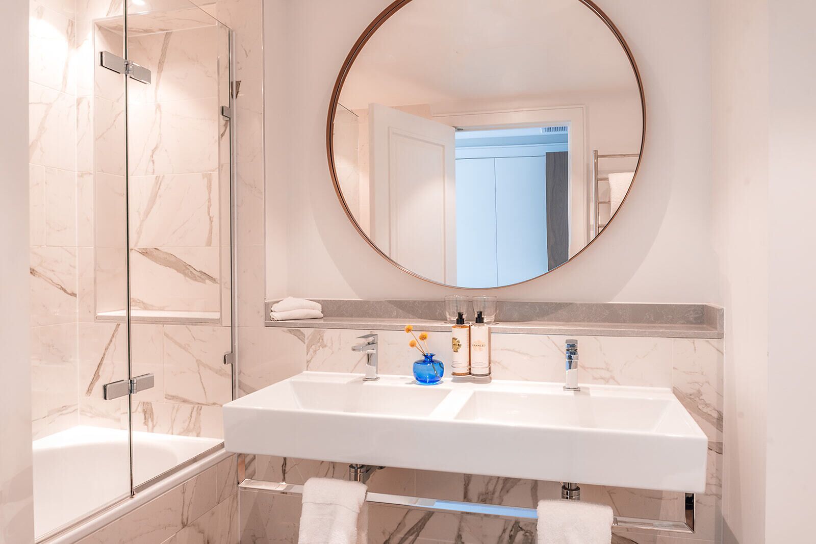 A modern bathroom with a glass shower enclosure, a white sink with a marble countertop, and a large round mirror.