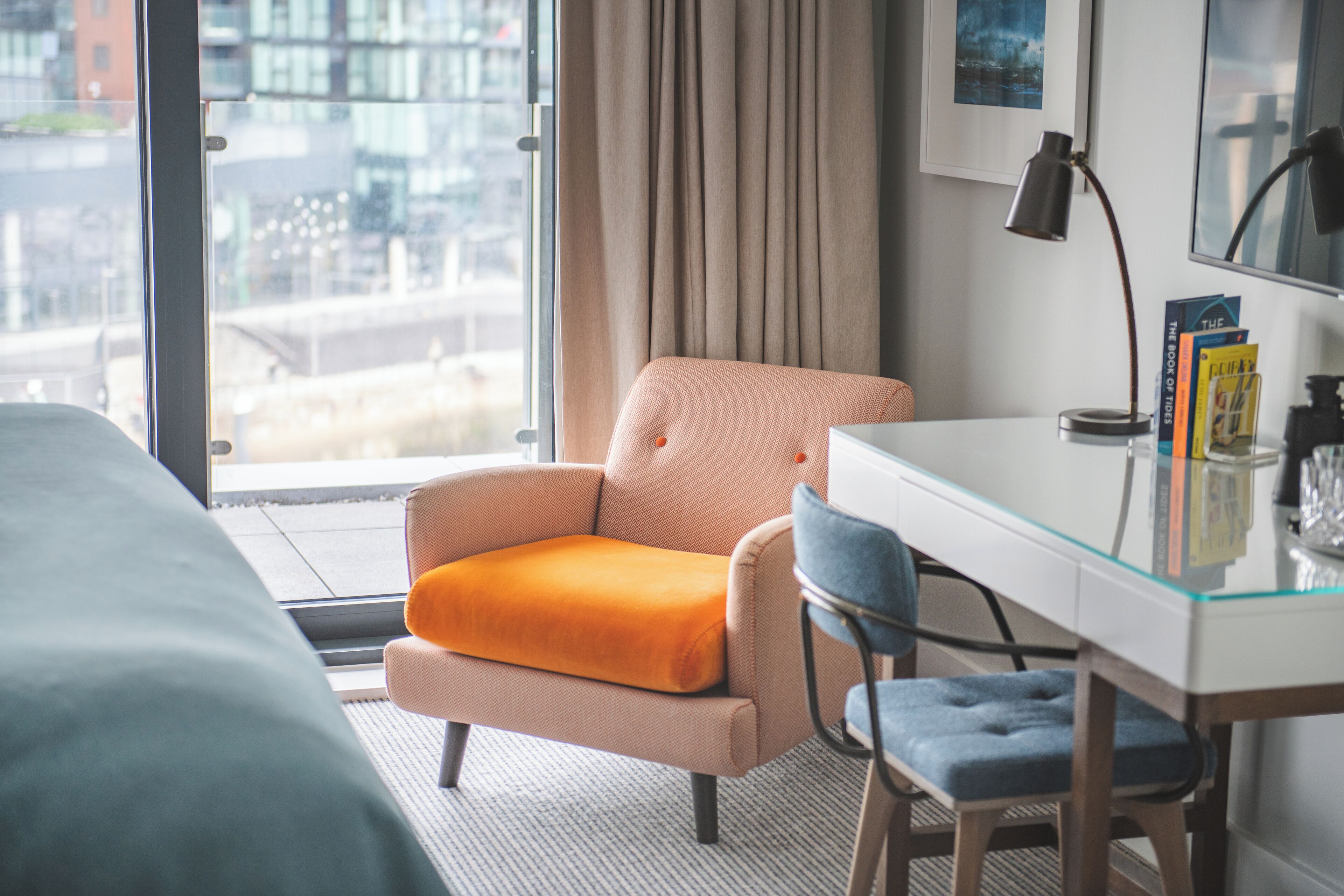 A hotel room with a bed, a desk with a lamp, a chair, and a view of the city through large windows.