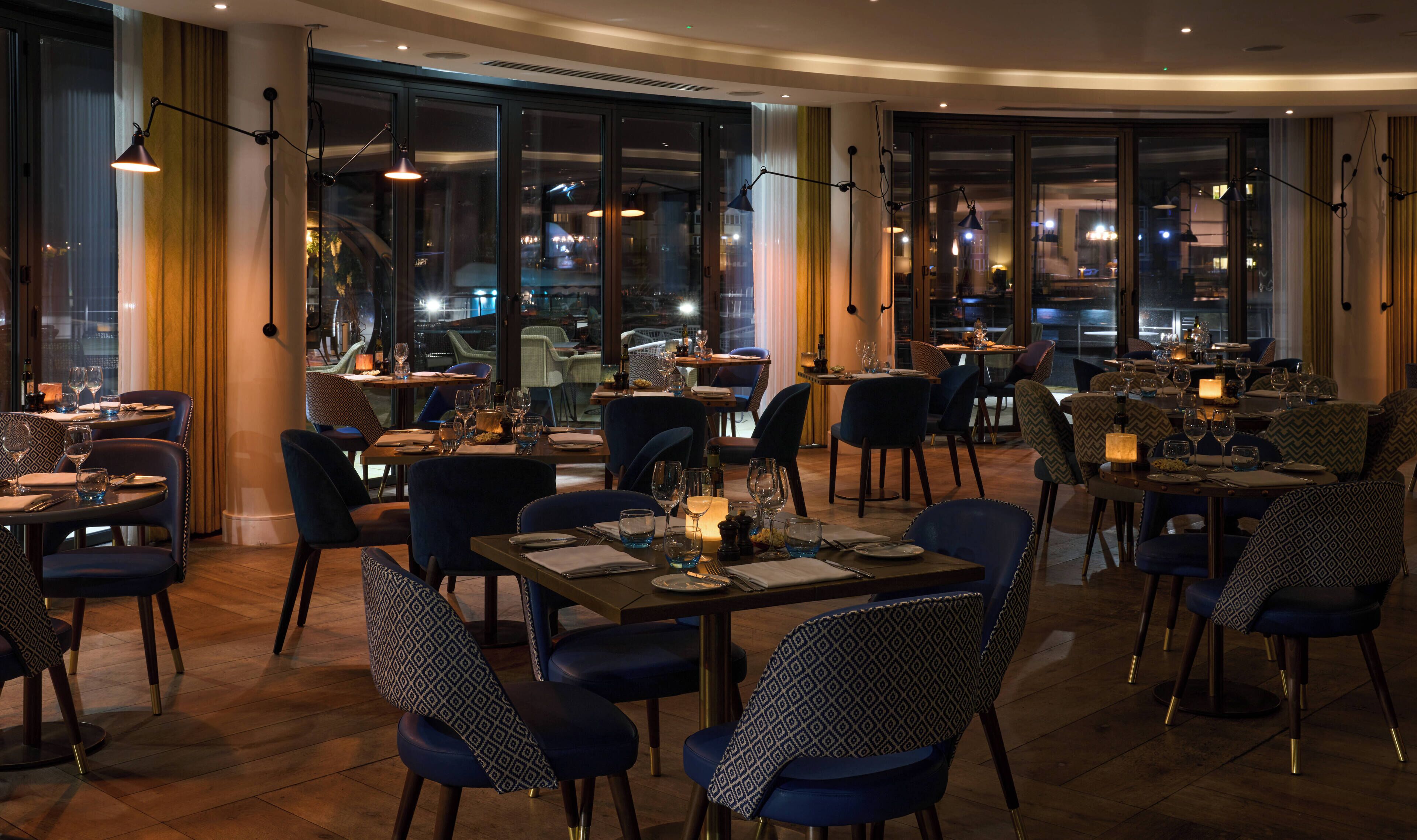 A restaurant with multiple tables and chairs, large windows, and pendant lighting.