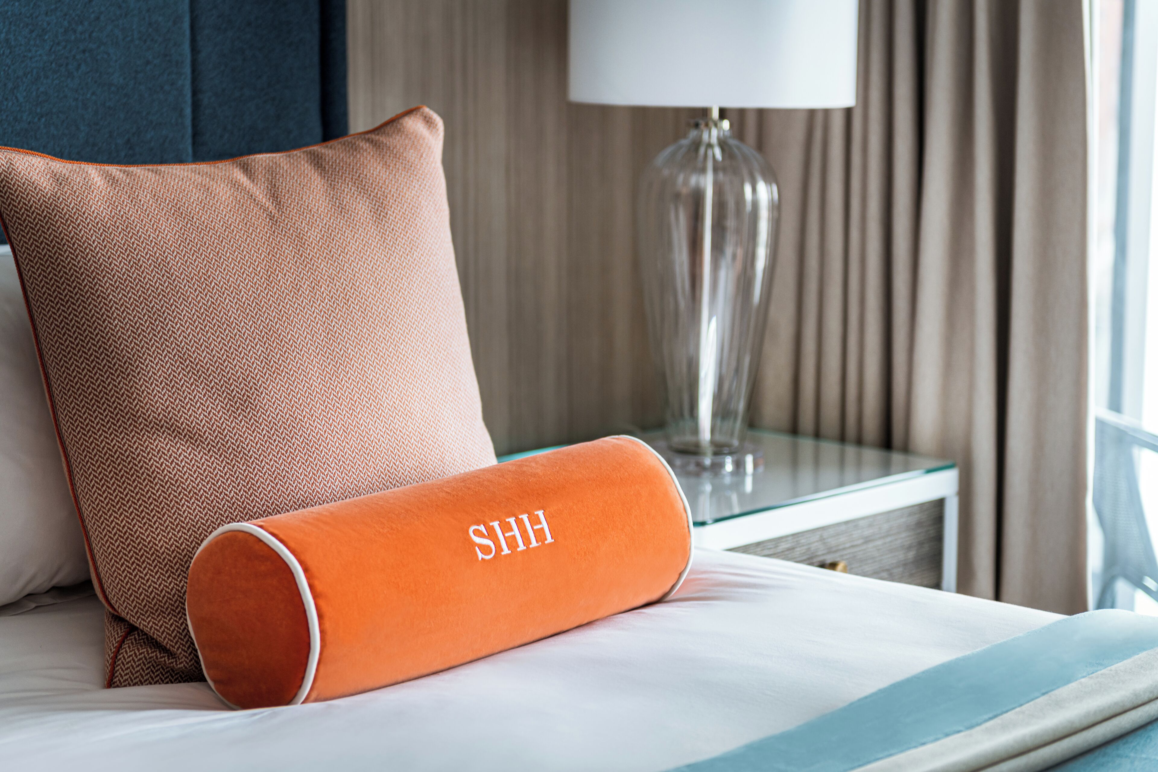 Un lit bien fait, recouvert d’une couvre-lit à rayures blanches et bleues, un oreiller orange orné du mot « CHUT » brodé dessus, et une lampe en verre transparent posée sur une table de chevet.