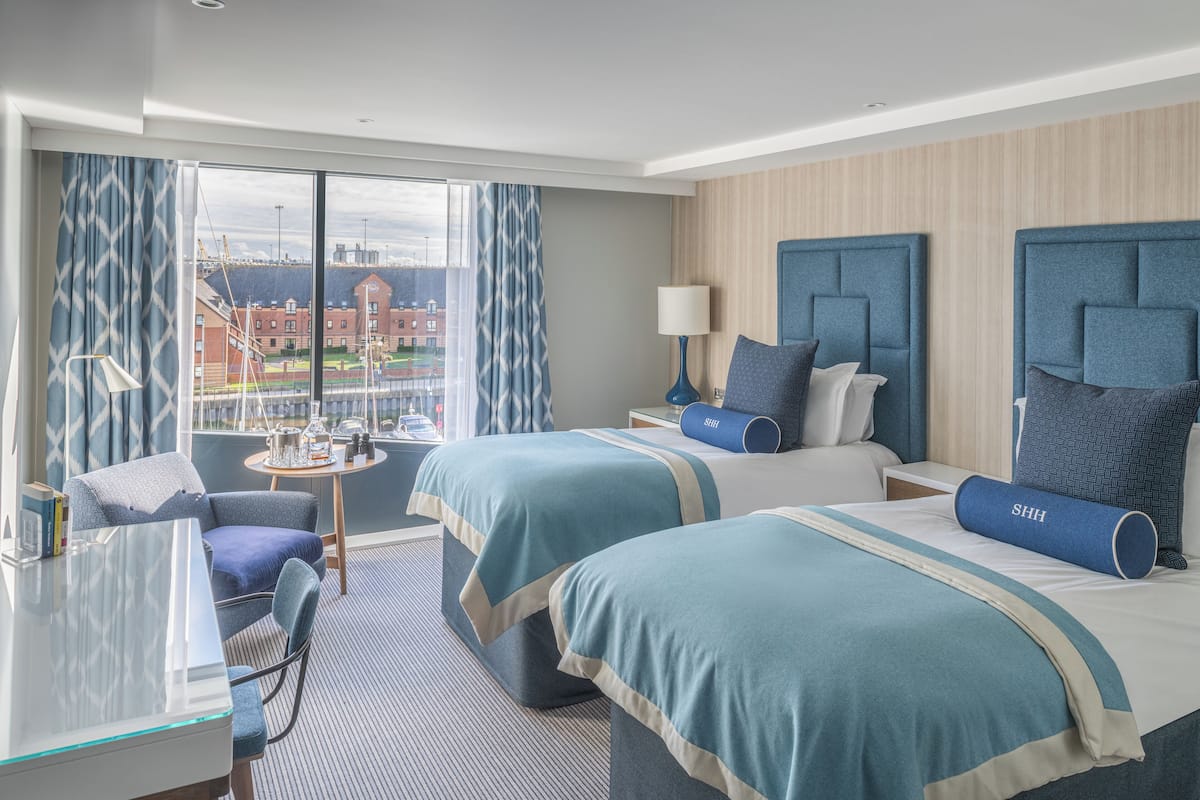 A hotel room with two beds, a desk, a chair, and a view of a cityscape.