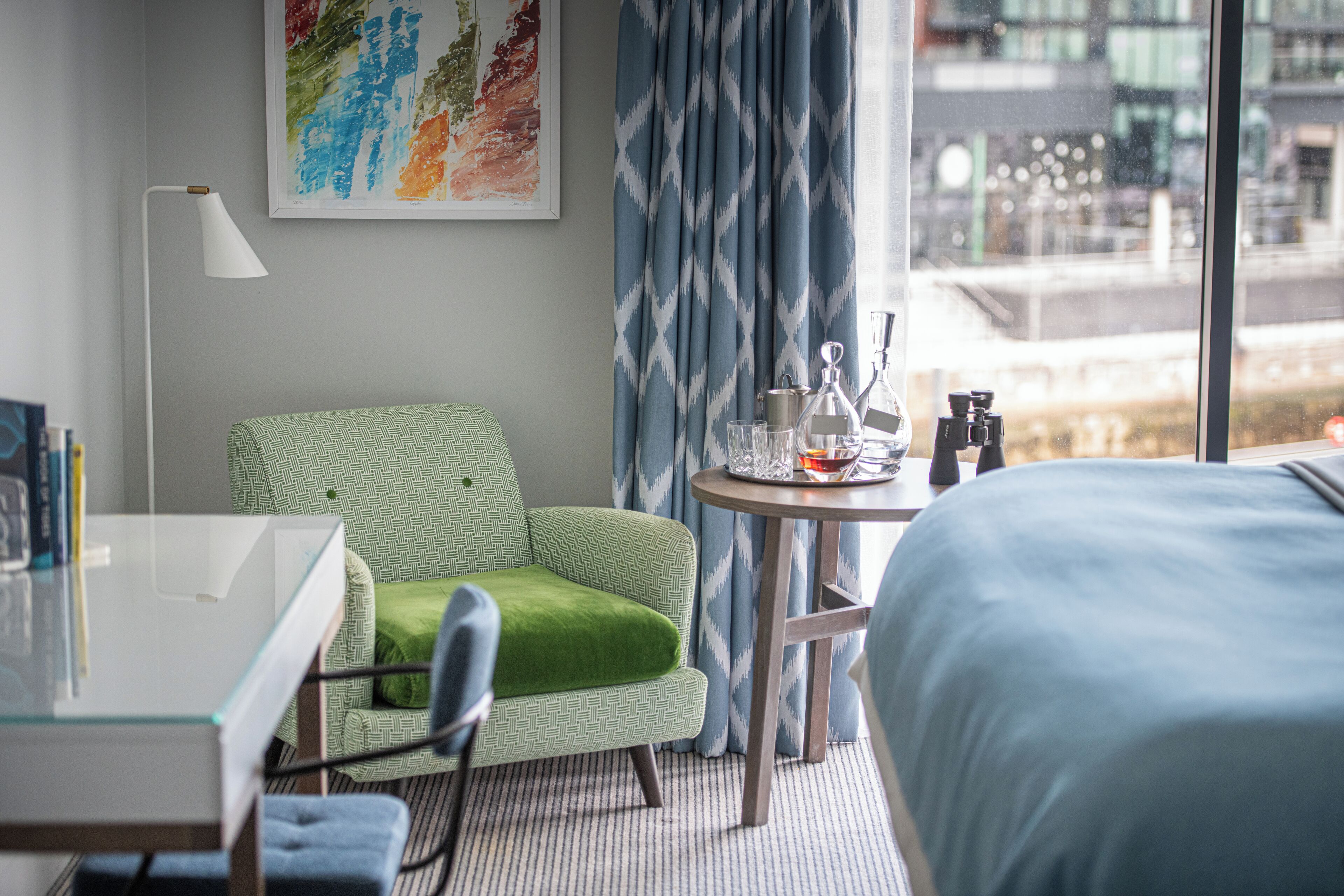Une chambre d’hôtel avec un lit, un fauteuil vert, une petite table avec une lampe et une fenêtre avec des rideaux.