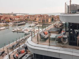 A marina with boats docked, a waterfront restaurant with outdoor seating, and residential buildings in the background.