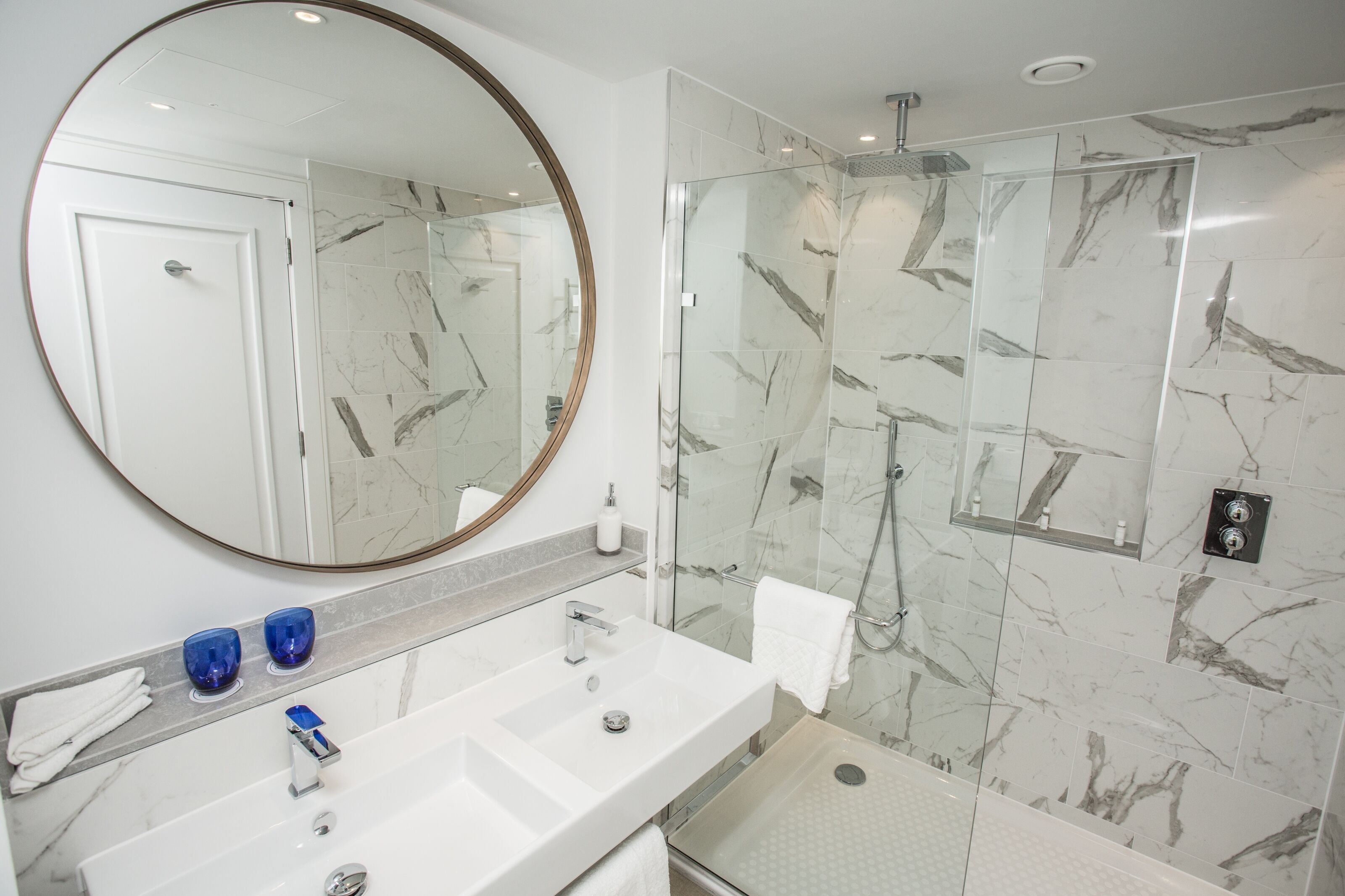 Une salle de bain moderne avec un grand miroir, deux vasques, une douche en verre et des murs à motifs de marbre.