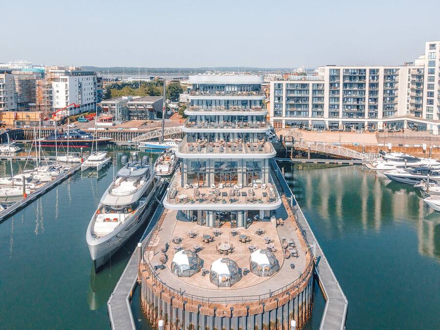 Un puerto deportivo frente al mar con un muelle para barcos de varios niveles y un restaurante con terraza.