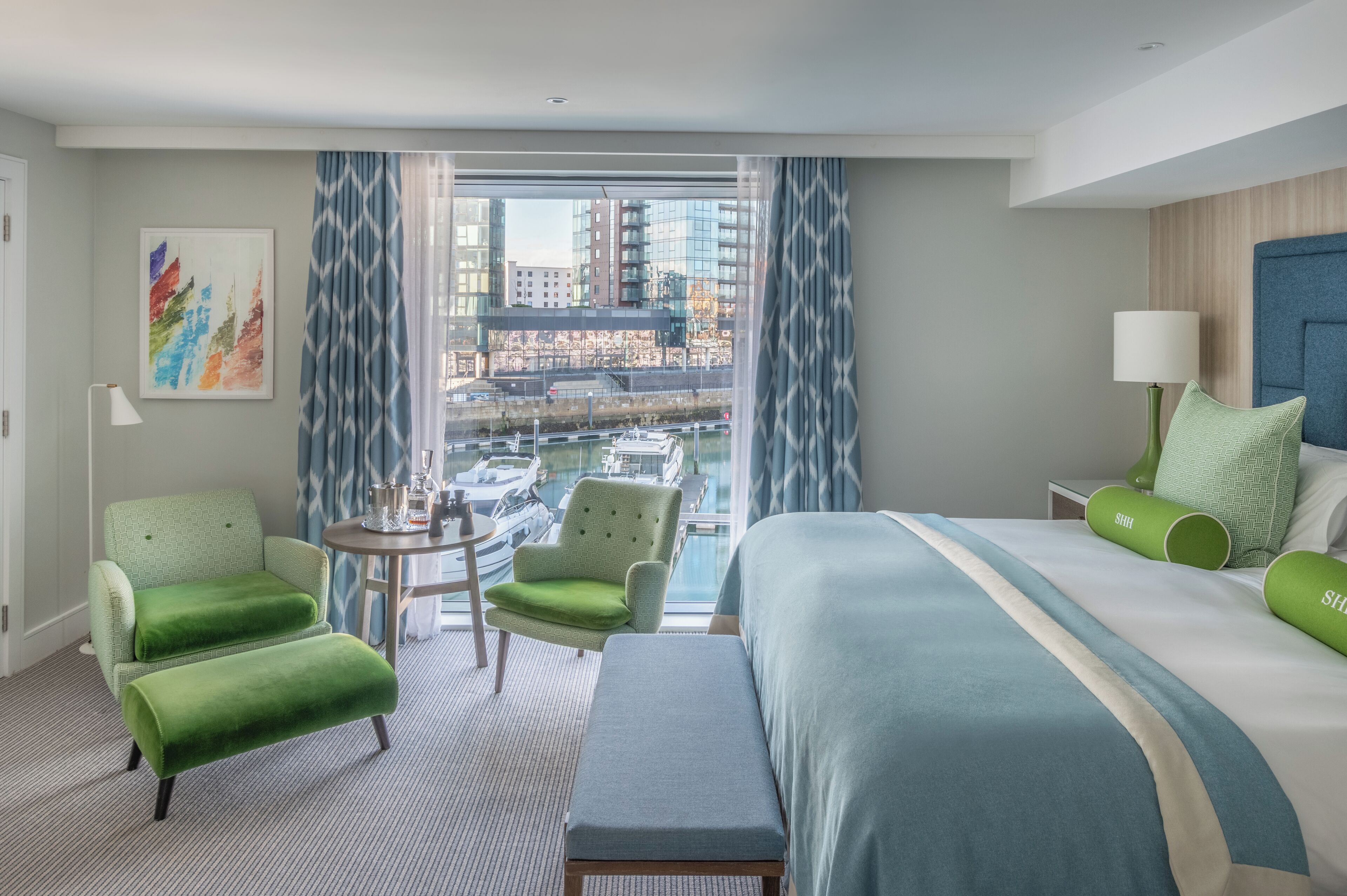 A hotel room with a large bed, two green armchairs, a small table, and a view of a cityscape and water through large windows.