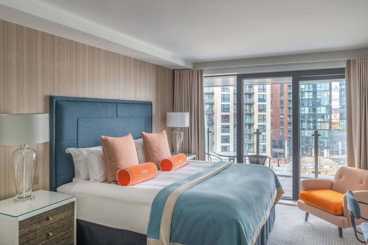 A hotel room with a large bed, a nightstand, a chair, and a view of the city through large windows.