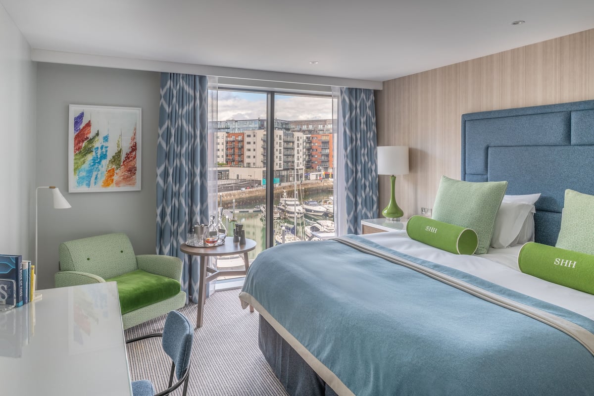 A hotel room with a large bed, a green sofa, a small table with a lamp, and a view of buildings through the window.