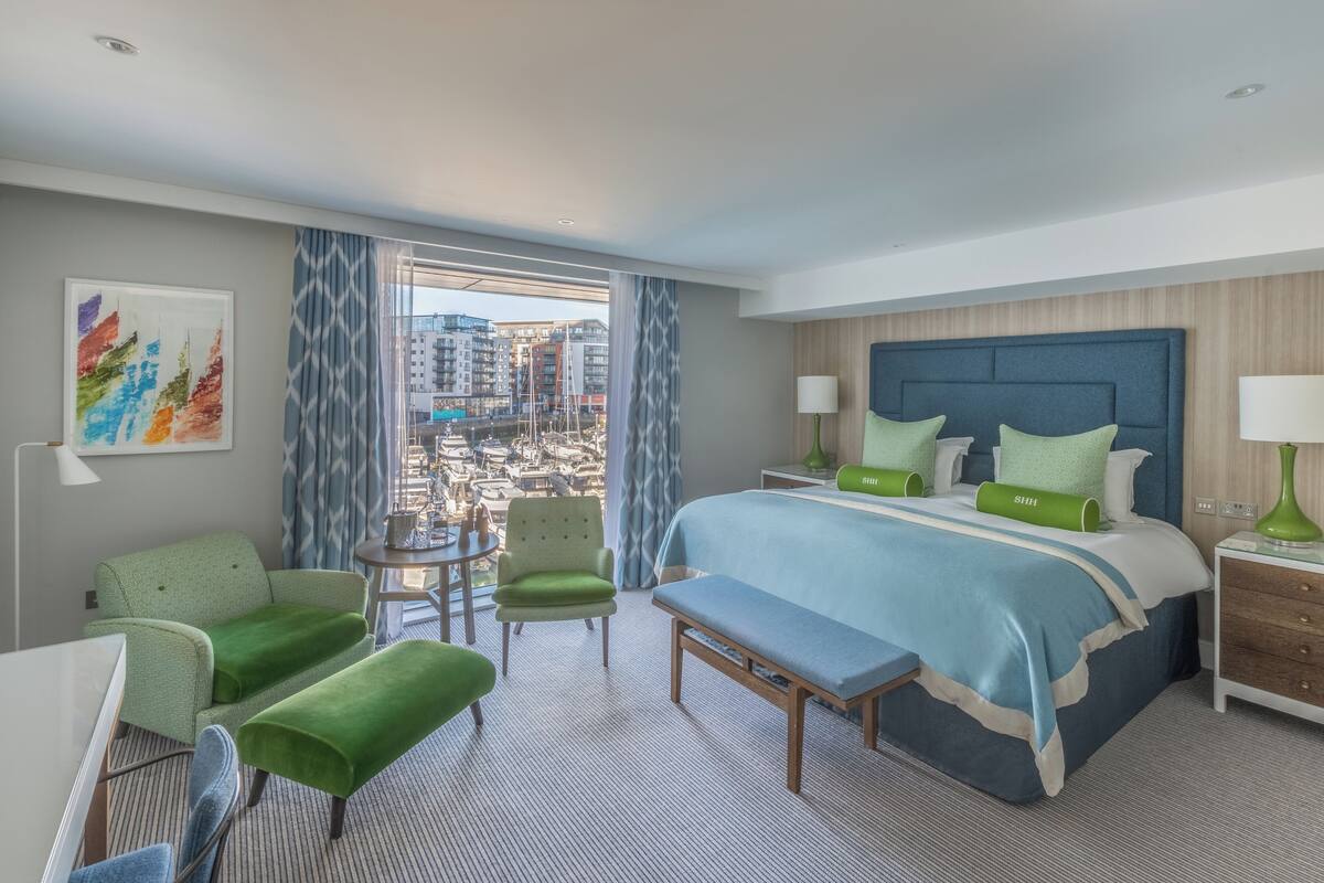 A hotel room with a large bed, green upholstered chairs, a small round table, and a view of a cityscape through large windows.