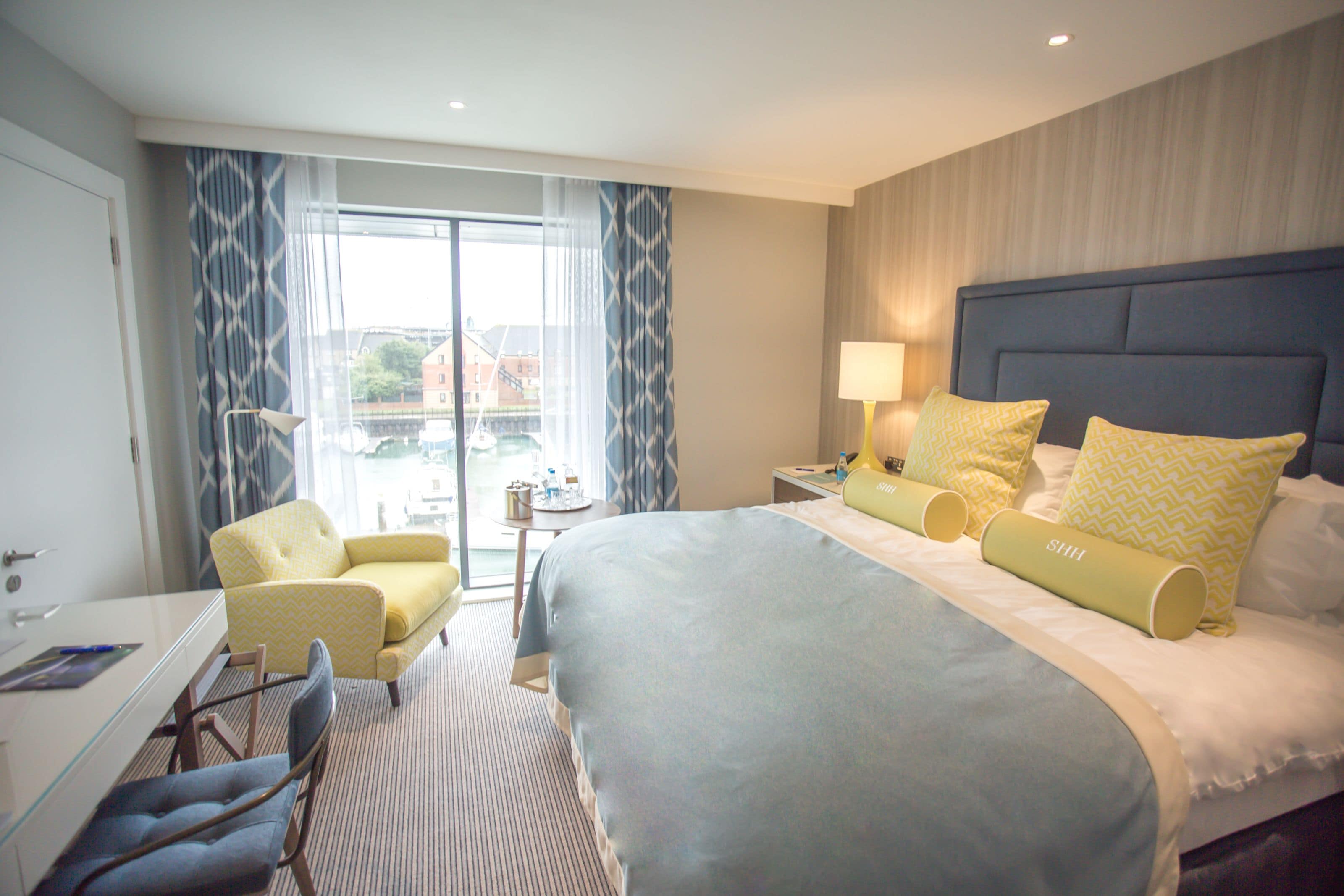A hotel room with a large bed, a desk, a chair, and a view of the outside.