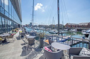 Una terraza con asientos al aire libre, con vista a un puerto deportivo con barcos y un paisaje urbano de fondo.