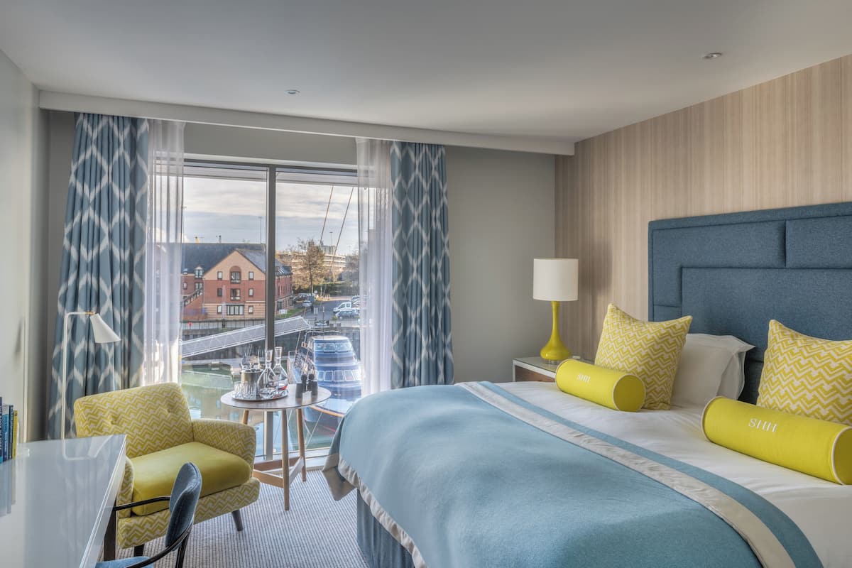 A hotel room with a large bed, a chair, a small table, and a view of the city through large windows.