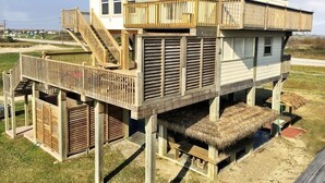 Una casa de playa de dos pisos con terraza de madera, balcón y techo de paja.