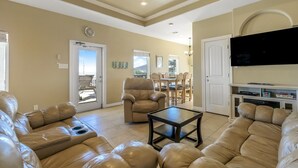 A living room with a beige sofa, a coffee table, and a television. There is a door leading to a balcony with a view of the mountains. - Parrot Head 3 Bedroom Home by RedAwning (Port Bolivar)