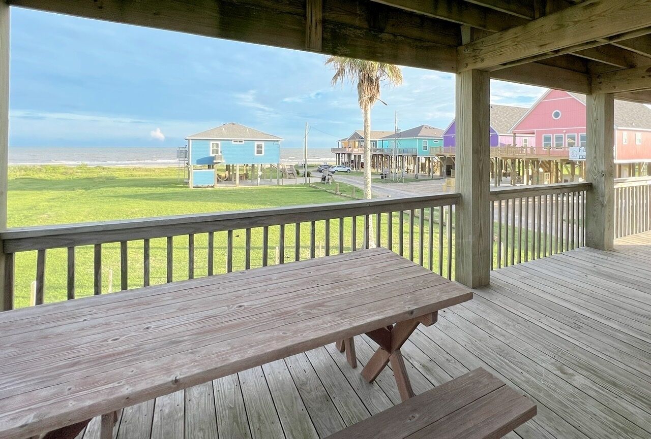 Une terrasse en bois avec une table de pique-nique, donnant sur une plage bordée de maisons et agrémentée d’un palmier.