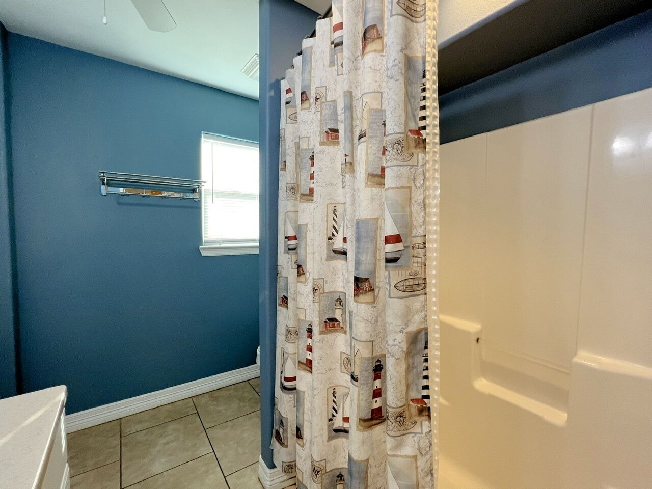 A bathroom with a blue wall, a window, a towel rack, and a shower curtain with nautical designs.