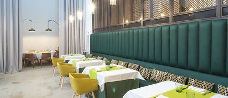 Un restaurant avec des banquettes recouvertes d’un tissu vert, des chaises jaunes et des tables dressées avec des nappes blanches et des sets de table verts.