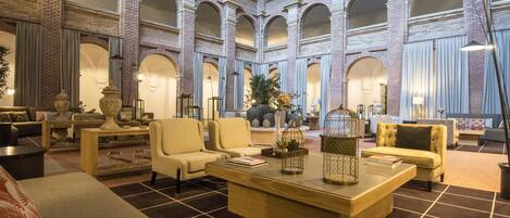 A spacious lobby with a high ceiling, featuring a central coffee table, seating area with sofas and armchairs, and a decorative plant centerpiece.