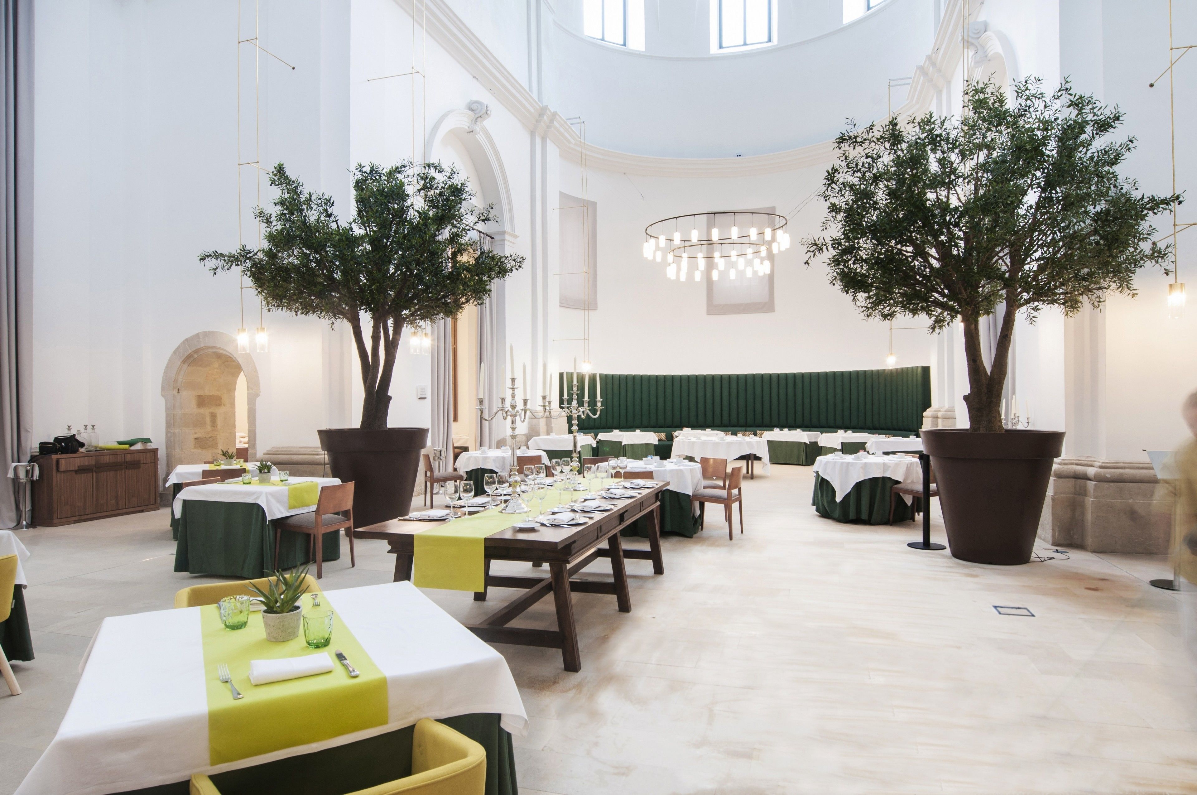 A spacious dining hall with high ceilings, large windows, and two potted trees. Tables are set for a meal, and there is a stage in the background.