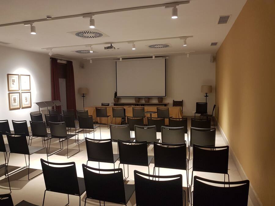 A conference room with a large screen, multiple rows of chairs, and ceiling lights.