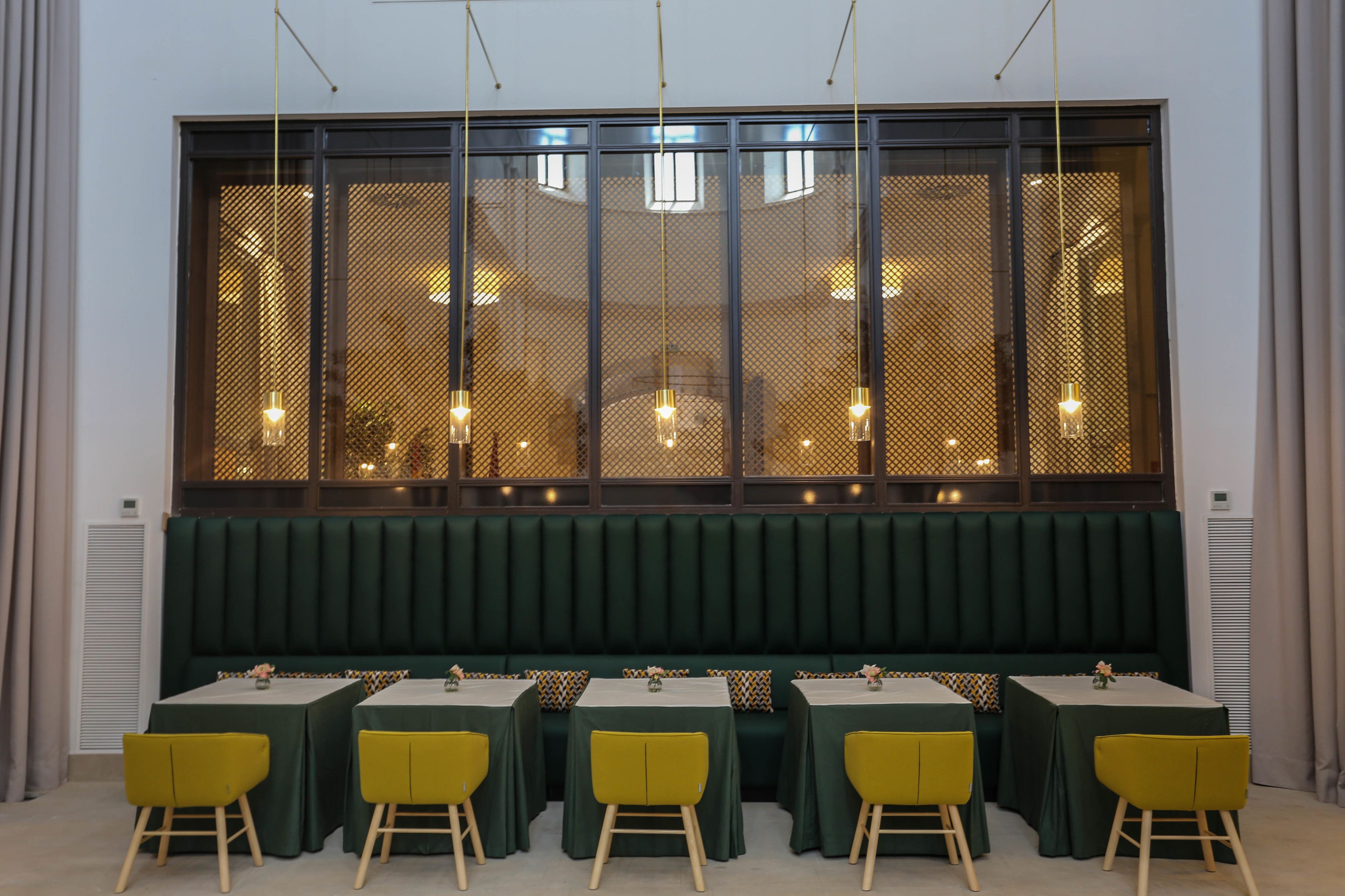 A restaurant with a row of tables and yellow chairs, a green upholstered bench, and a large window with hanging lights.